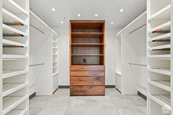 Walk-in closet featuring white custom shelving units, central wood-finish cabinet with built-in drawers and open shelving, integrated hanging rods, and large-format tile flooring