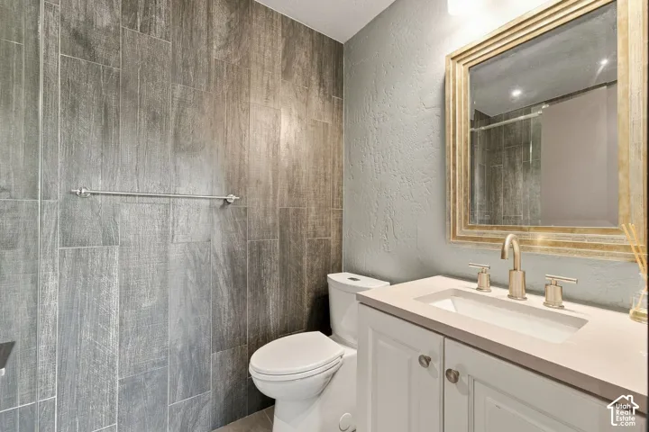 Bathroom featuring floor-to-ceiling wood-finish tiled walls, white shaker-style vanity cabinet with a stone-composite countertop, under-mount sink, brushed-finish widespread faucet, and a wall-mounted framed mirror