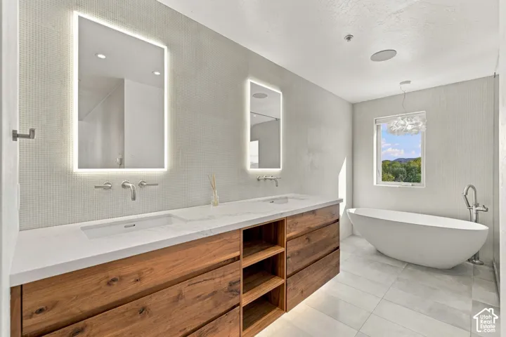 Dual-vanity with wood cabinetry, light-toned countertop, wall-mounted faucets, integrated LED-backlit mirrors, and freestanding soaking tub