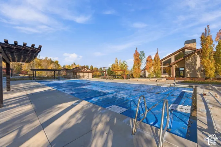 Swimming pool with metal handrails and concrete decking, featuring overhead wooden pergolas and a stone-accented exterior building