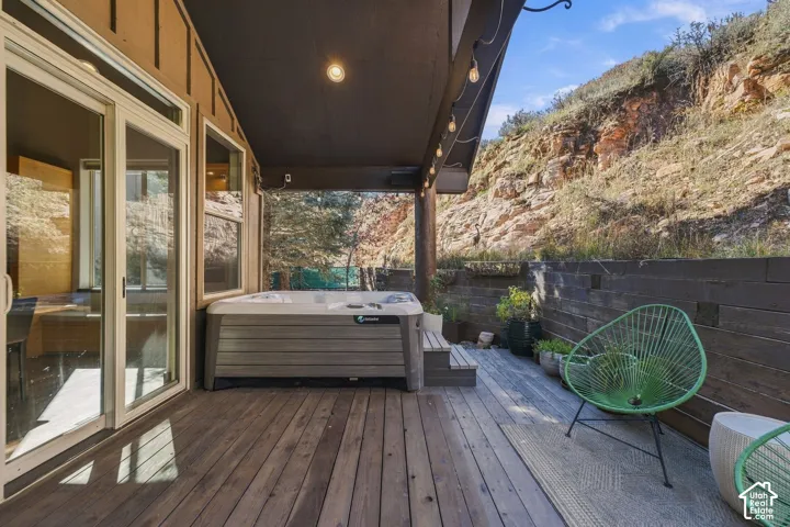 Covered outdoor deck featuring wood-finish decking, built-in hot tub, horizontal wood privacy fencing, and recessed ceiling lighting with mountainous rock terrain backdrop