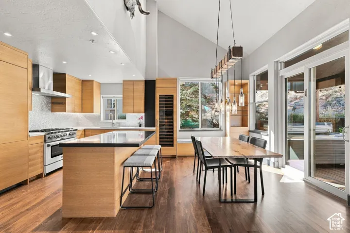 Kitchen featuring wood-finish cabinetry, a large center island with a dark countertop, integrated wine refrigeration, wood-finish flooring, and vaulted ceilings with suspended feature lighting