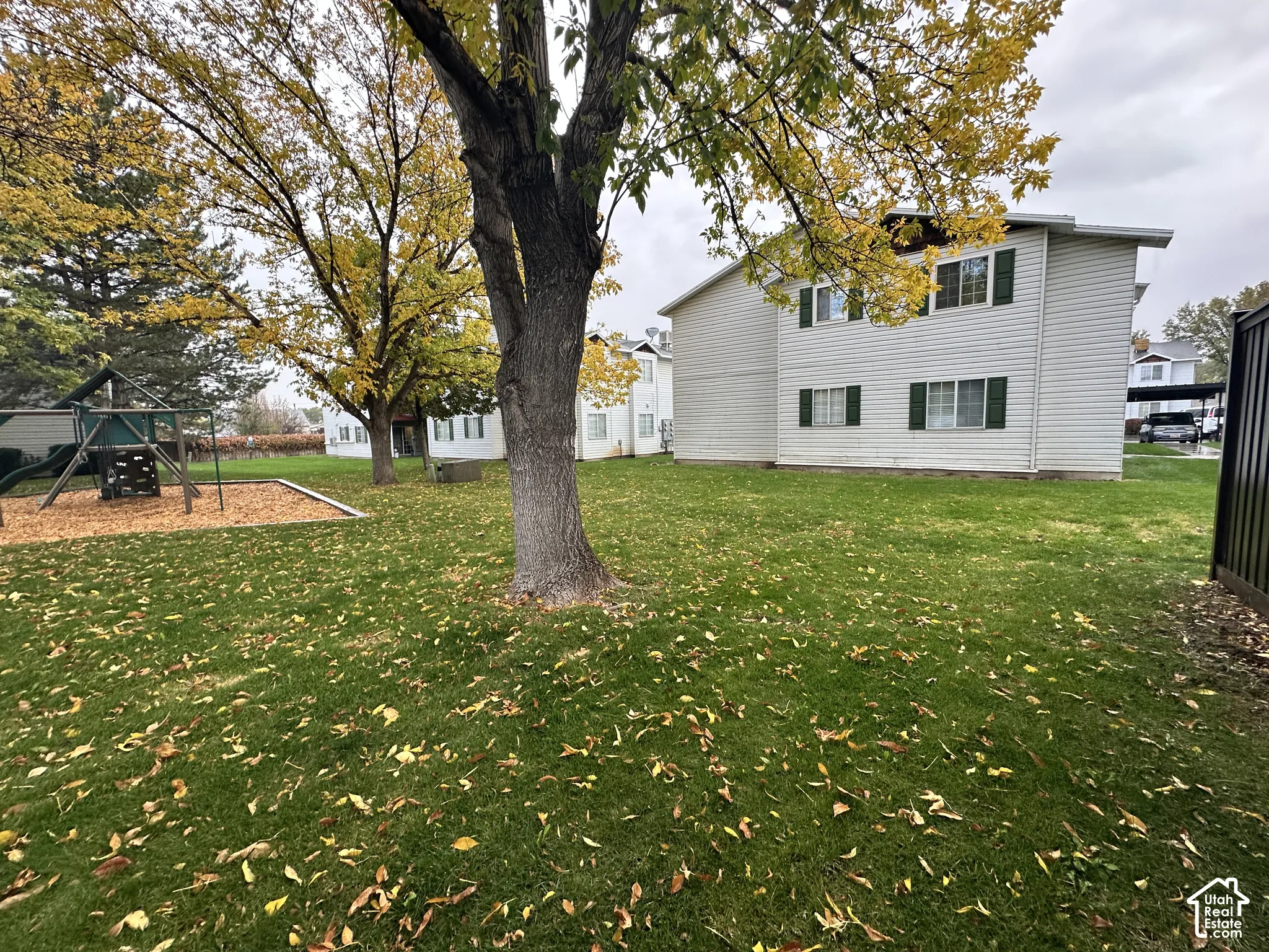 Exterior grounds featuring a multi-story building with white vinyl siding, green shutters, mature trees, and a ground-level play structure with a wood-chip base