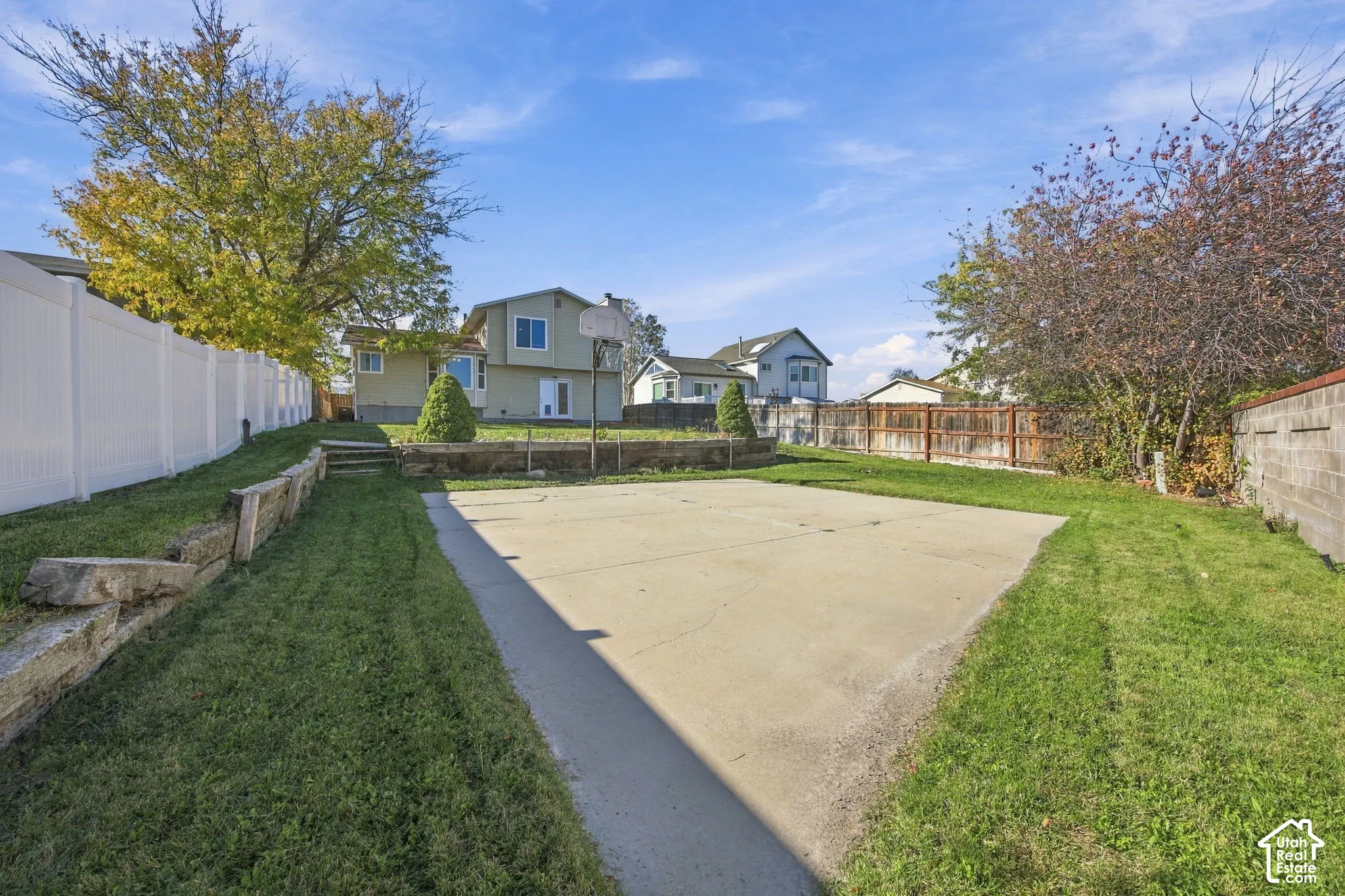 Expansive backyard featuring a concrete sport court, tiered timber retaining walls, mature landscaping, and vinyl perimeter fencing