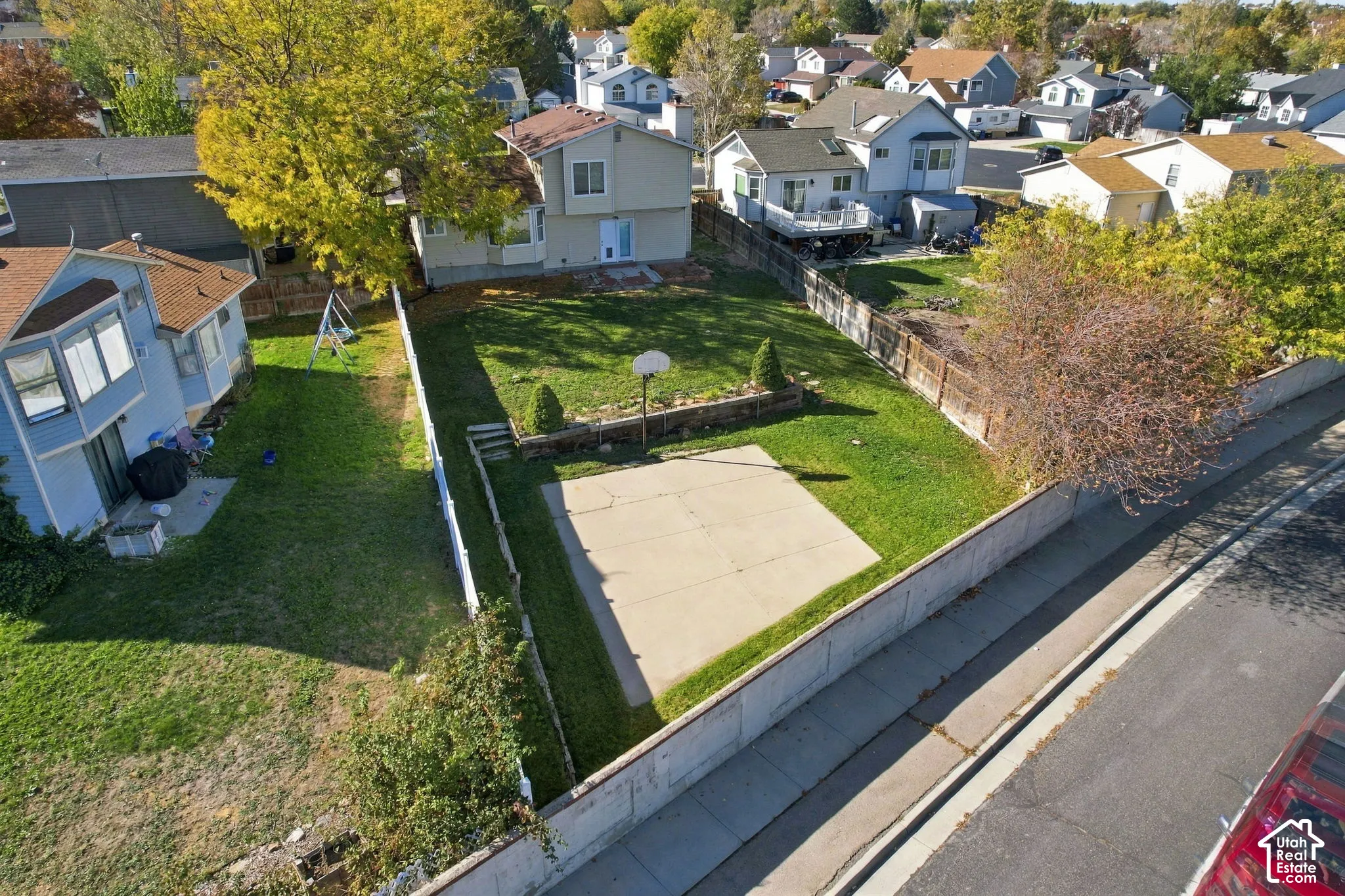 Fenced backyard featuring a concrete sport court, tiered stone retaining wall, mature landscaping, and an expansive lawn