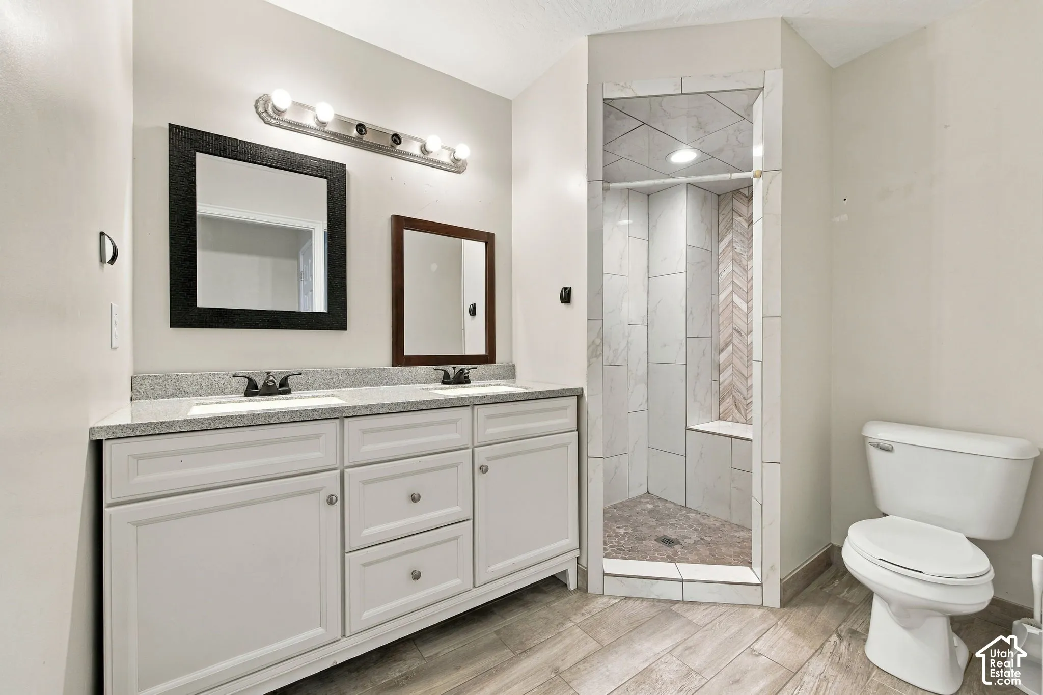 Dual-sink vanity with stone countertop and white cabinetry, accompanied by a walk-in shower featuring herringbone-patterned tile accents and wood-finish flooring