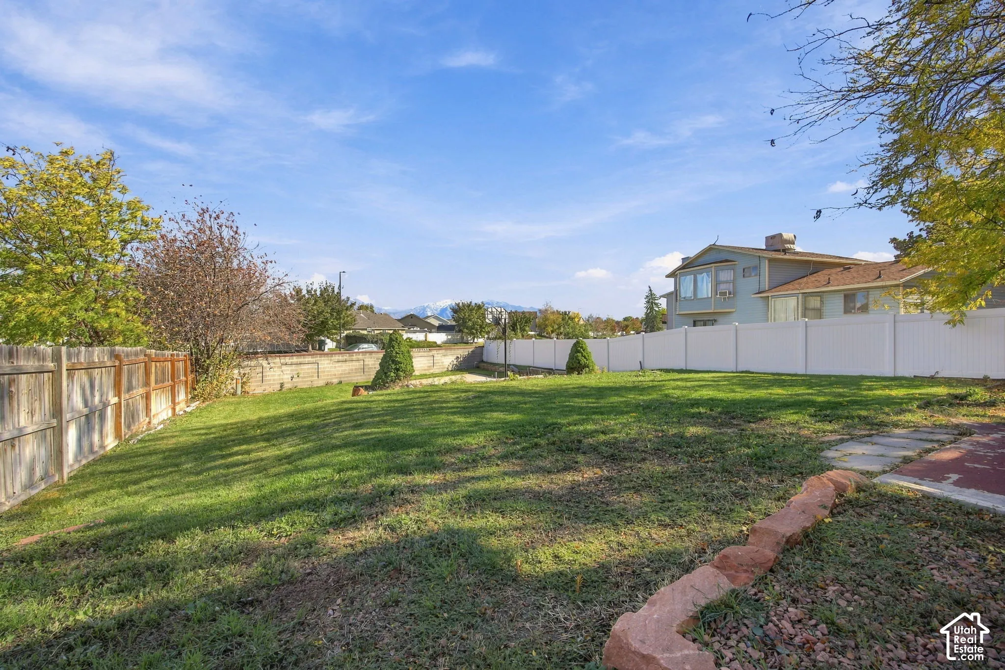 Expansive backyard featuring a large grass lawn, white vinyl fencing, a natural stone retaining wall, and mature trees with mountain backdrop