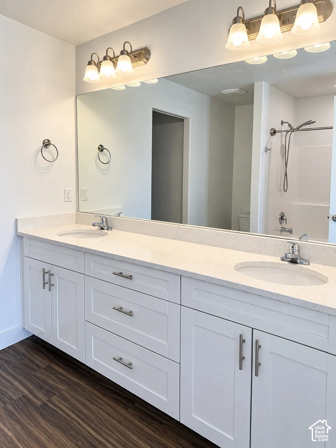 Double vanity featuring a light-toned solid surface countertop, white shaker-style cabinetry, metallic hardware, an expansive wall-mounted mirror, and wood-finish flooring