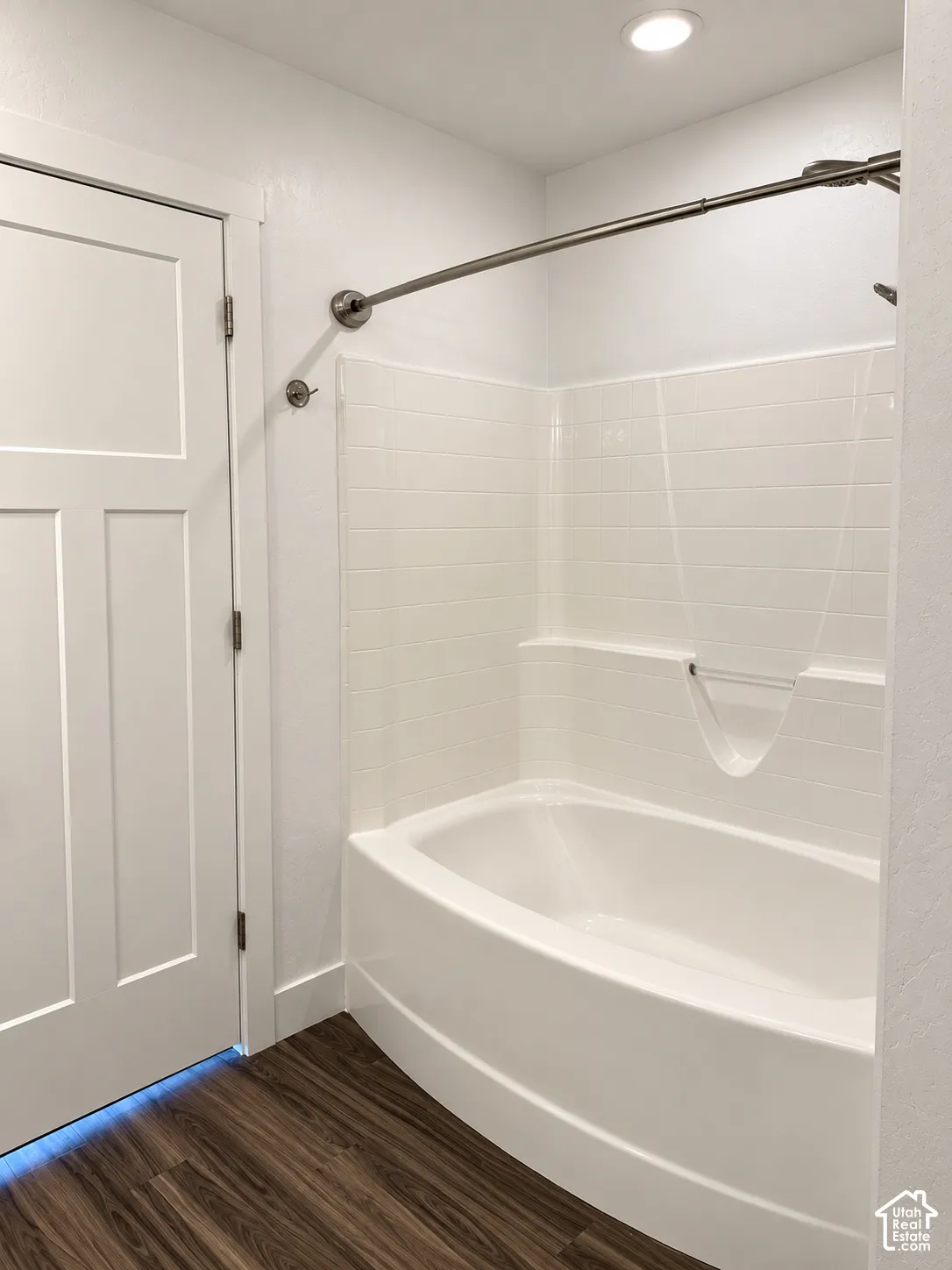 Curved acrylic bathtub with integrated tile-pattern surround, polished nickel shower rod, and wood-finish flooring