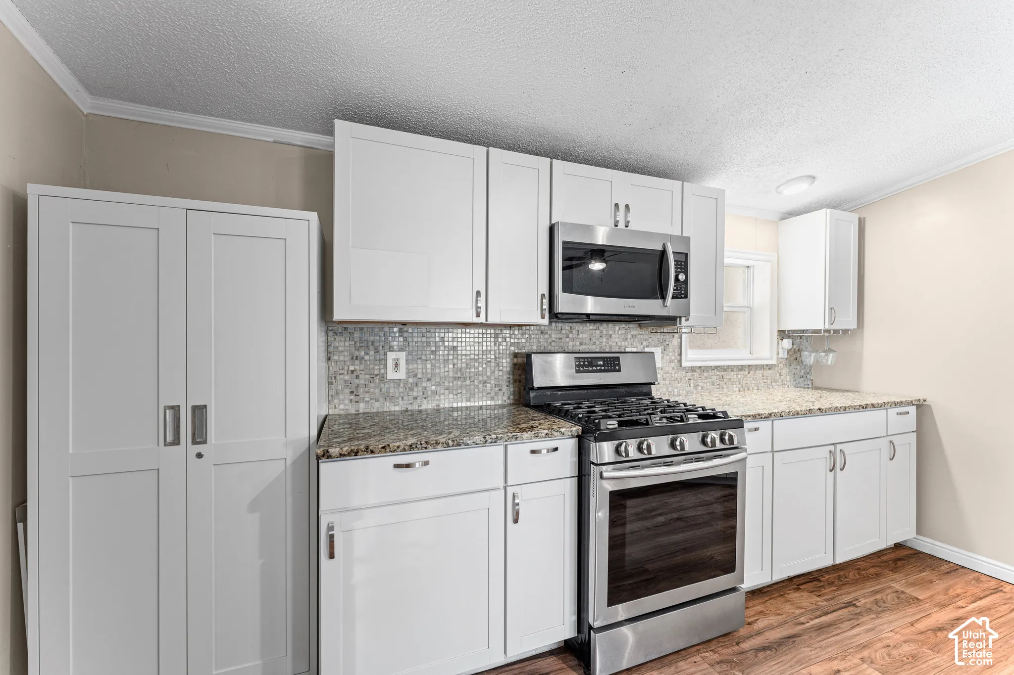 Kitchen featuring white shaker-style cabinetry, stone-finish countertops, stainless steel gas range and microwave, mosaic tile backsplash, and wood-finish flooring