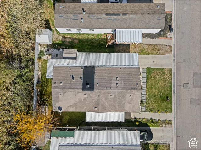 Aerial view featuring a paved driveway, metal roofing, structured hardscaping, and mature perimeter trees