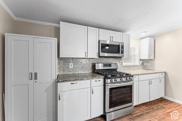 Kitchen featuring white shaker-style cabinetry, stone-finish countertops, stainless steel gas range and microwave, mosaic tile backsplash, and wood-finish flooring