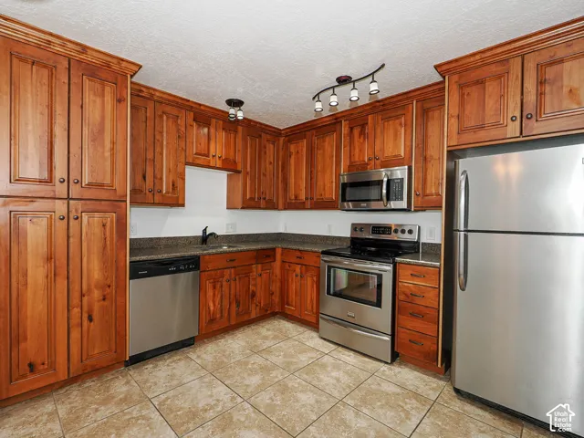 Kitchen featuring stained wood cabinetry, stone-finish countertops, tile flooring, stainless steel appliances, and multi-bulb track lighting