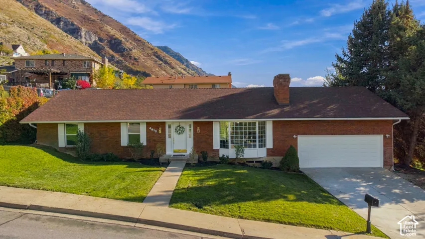 Single-level brick exterior featuring a multi-pane bay window, an attached two-car garage, an asphalt shingle roof, and a prominent brick chimney, set against a mountainous backdrop