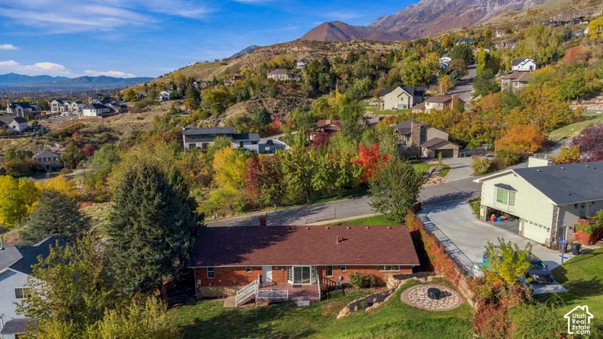 Aerial perspective featuring a brick-exterior residence, a rear wooden deck, a circular stone hardscape feature, mature trees, and a mountain backdrop