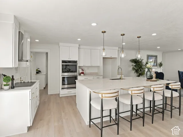 Kitchen featuring white shaker-style cabinetry, quartz countertops, a central island with an integrated sink, built-in double wall ovens, and wood-finish flooring