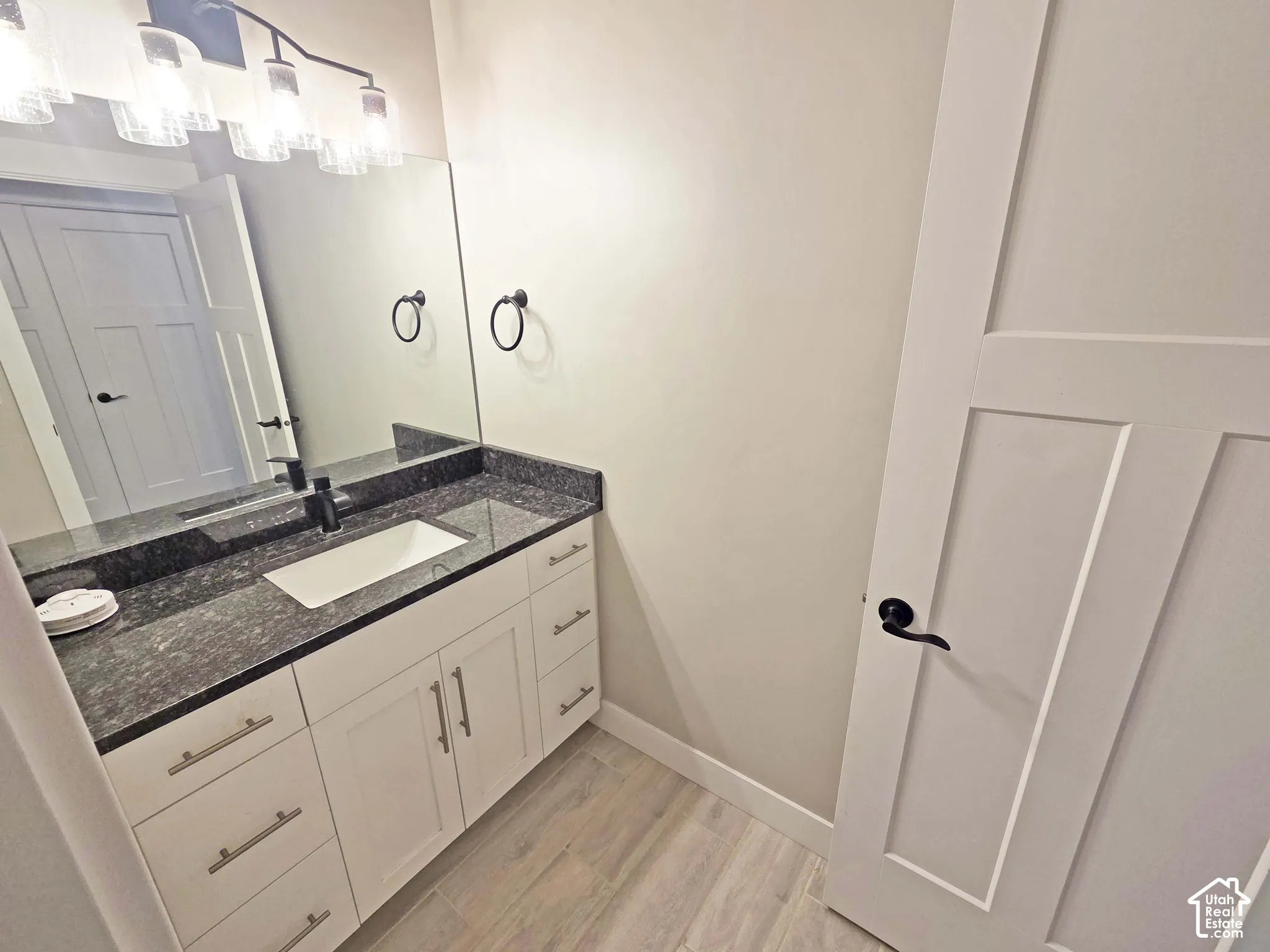 White cabinetry vanity featuring a stone countertop, integrated rectangular sink, matte black faucet, and hardware, complemented by wood-finish flooring and a shaker-style interior door