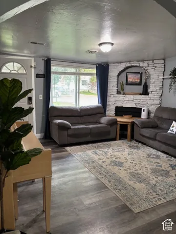 Living area featuring wood-finish flooring, a white stacked stone fireplace with an arched hearth, and a large multi-pane window