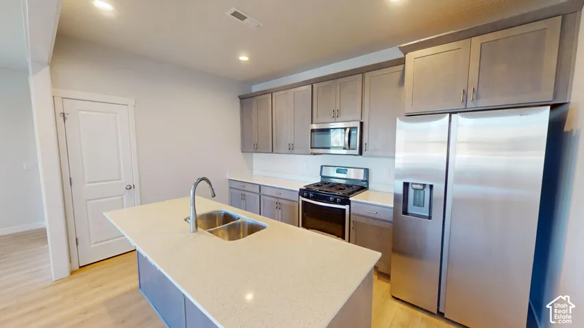 Kitchen featuring gray cabinetry, light-toned countertops, a center island with a dual-basin sink, and stainless steel appliances including a gas range and refrigerator