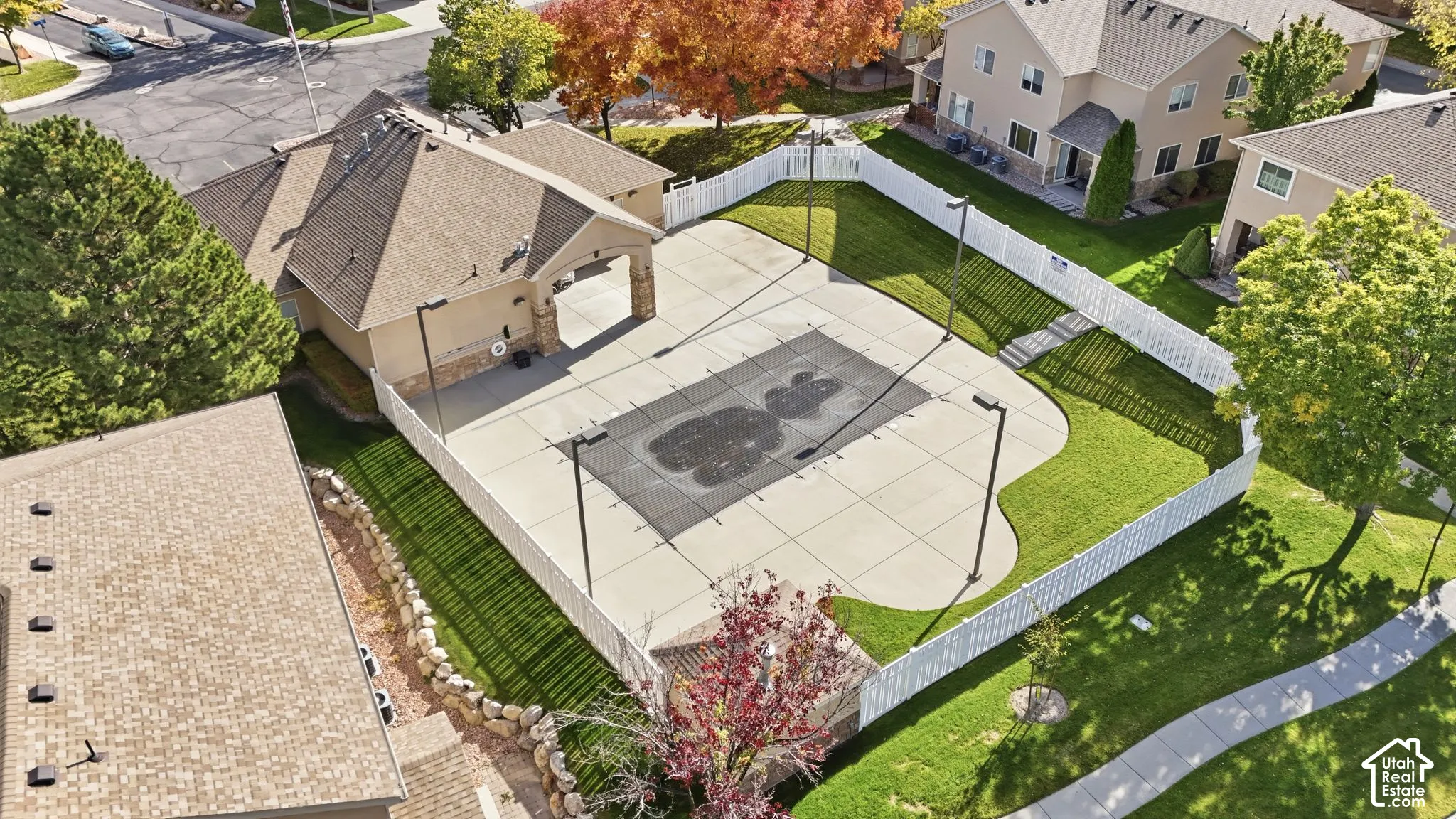 Fenced recreation area featuring a concrete sports court, high-mast lighting, stone-accented pavilion, and surrounding lawn