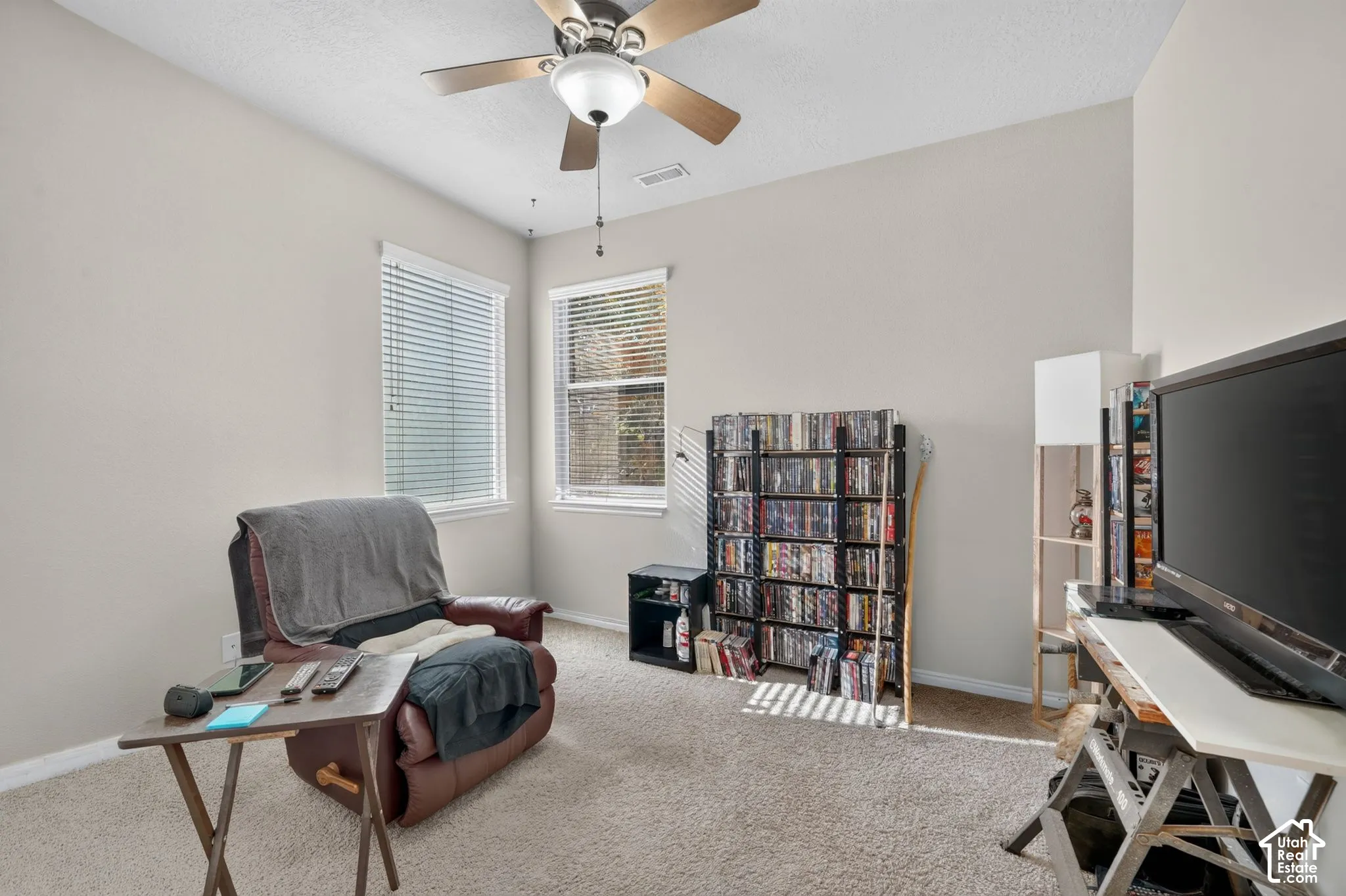 Room featuring neutral wall tones, plush wall-to-wall carpeting, two dual-pane windows with horizontal blinds, and a central ceiling fan with integrated lighting
