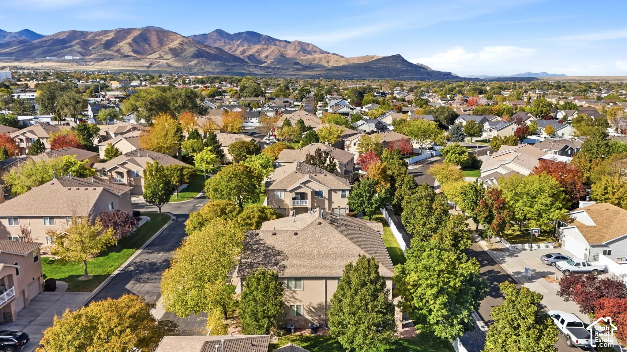 Aerial perspective of a multi-level residential neighborhood featuring asphalt paved streets, mature deciduous trees, a backdrop of mountain ranges, and hip-roof residential structures