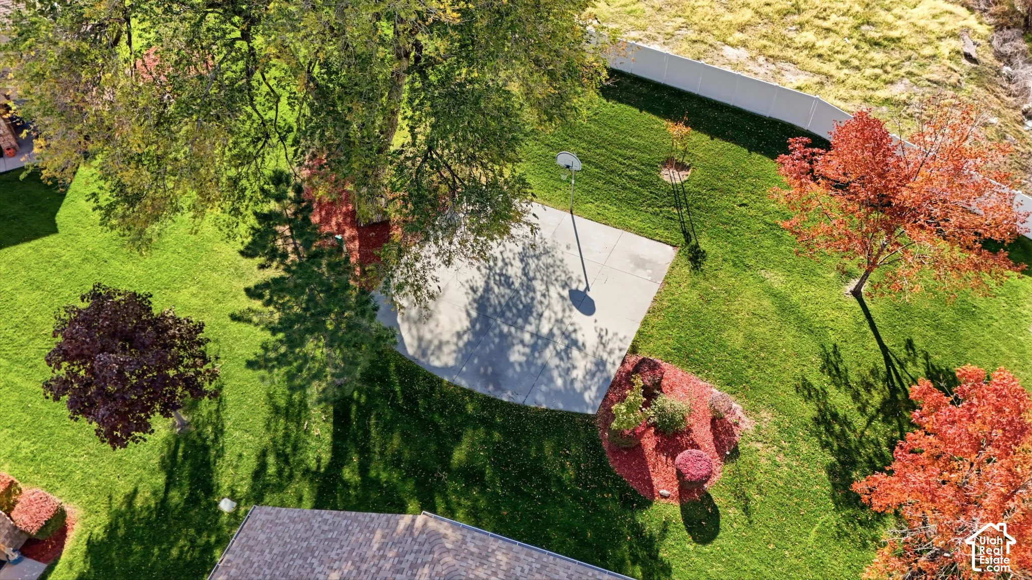 Expansive backyard featuring a concrete sport court, mature trees, landscaped planting beds, and white vinyl perimeter fencing