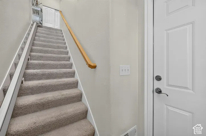 Staircase featuring neutral carpeted treads, a wooden handrail, and an attached stair lift system, accompanied by a paneled door with a metallic lever handle