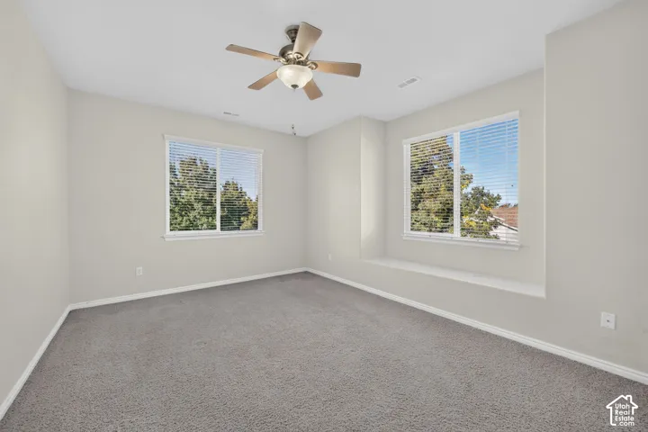 Neutral-toned interior featuring plush carpeting, a built-in window seat, two windows with horizontal blinds, and a ceiling fan with integrated lighting