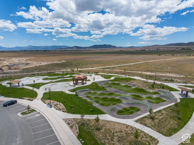 Recreational park area featuring a paved pump track, concrete pathways, covered gazebos, manicured lawn, and mountain scenery