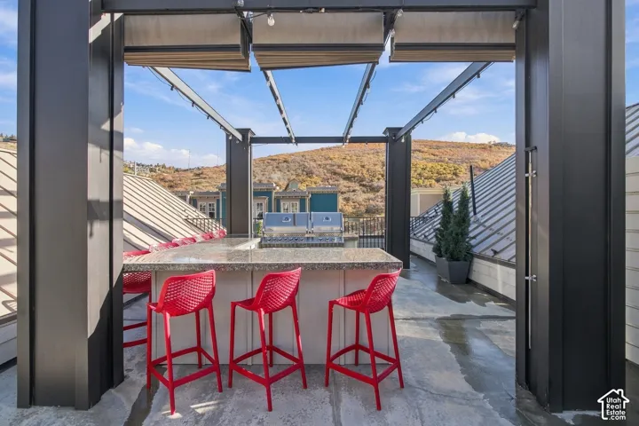 Rooftop terrace featuring a built-in outdoor kitchen with stone-finish countertops, a stainless steel grill, a metal-framed pergola with retractable canopy, and mountain scenery