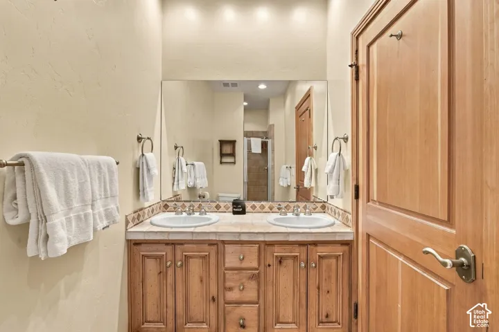 Dual-sink vanity featuring custom wood cabinetry, tiled countertop, decorative backsplash inlay, and recessed lighting