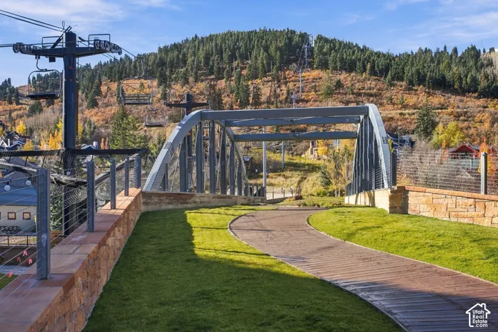 Pedestrian path featuring a curved wood-plank walkway, manicured grass borders, stacked-stone retaining walls, and a structural steel truss bridge