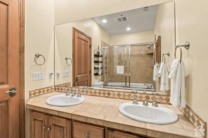 Dual-sink vanity featuring tile countertops with decorative diamond-patterned backsplash, solid wood cabinetry, a large wall-mounted mirror, and a tile-surround shower with sliding glass doors