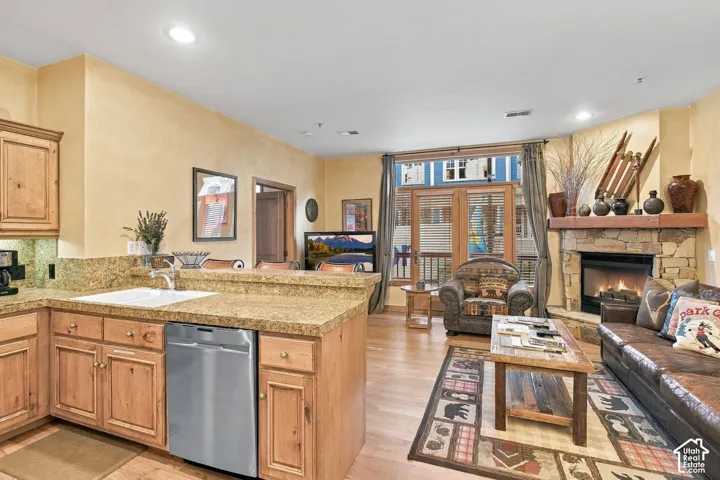 Natural stone fireplace with a thick timber mantel, custom wood cabinetry, stone countertops, stainless steel dishwasher, and wood-finish flooring