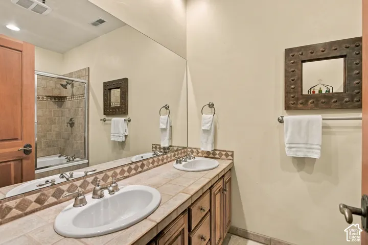 Full-width vanity featuring dual white undermount sinks, tile countertop with decorative accent backsplash, wood cabinetry, and large-format wall mirror