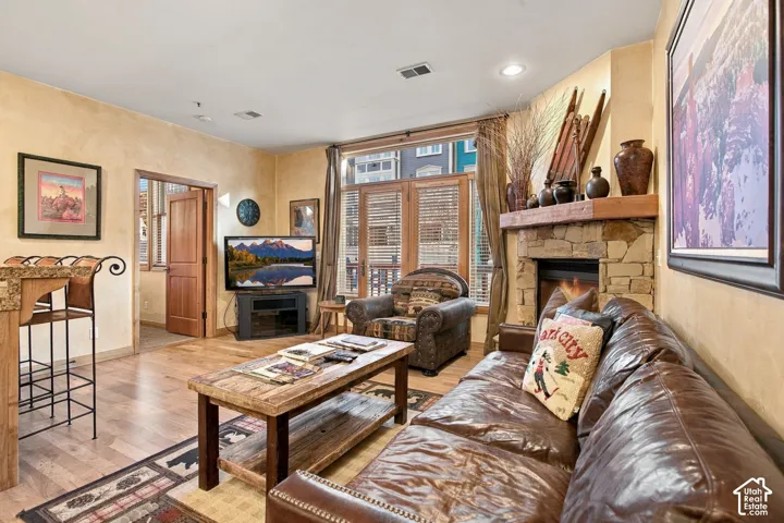 Living area featuring wood-finish flooring, a stone-surround fireplace with a timber mantel, and large windows with integrated shutters
