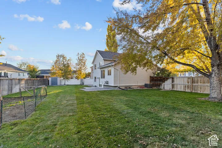 Expansive backyard featuring a large grass lawn, perimeter wood fencing, mature trees, a concrete patio, and a designated gardening area with wire enclosure
