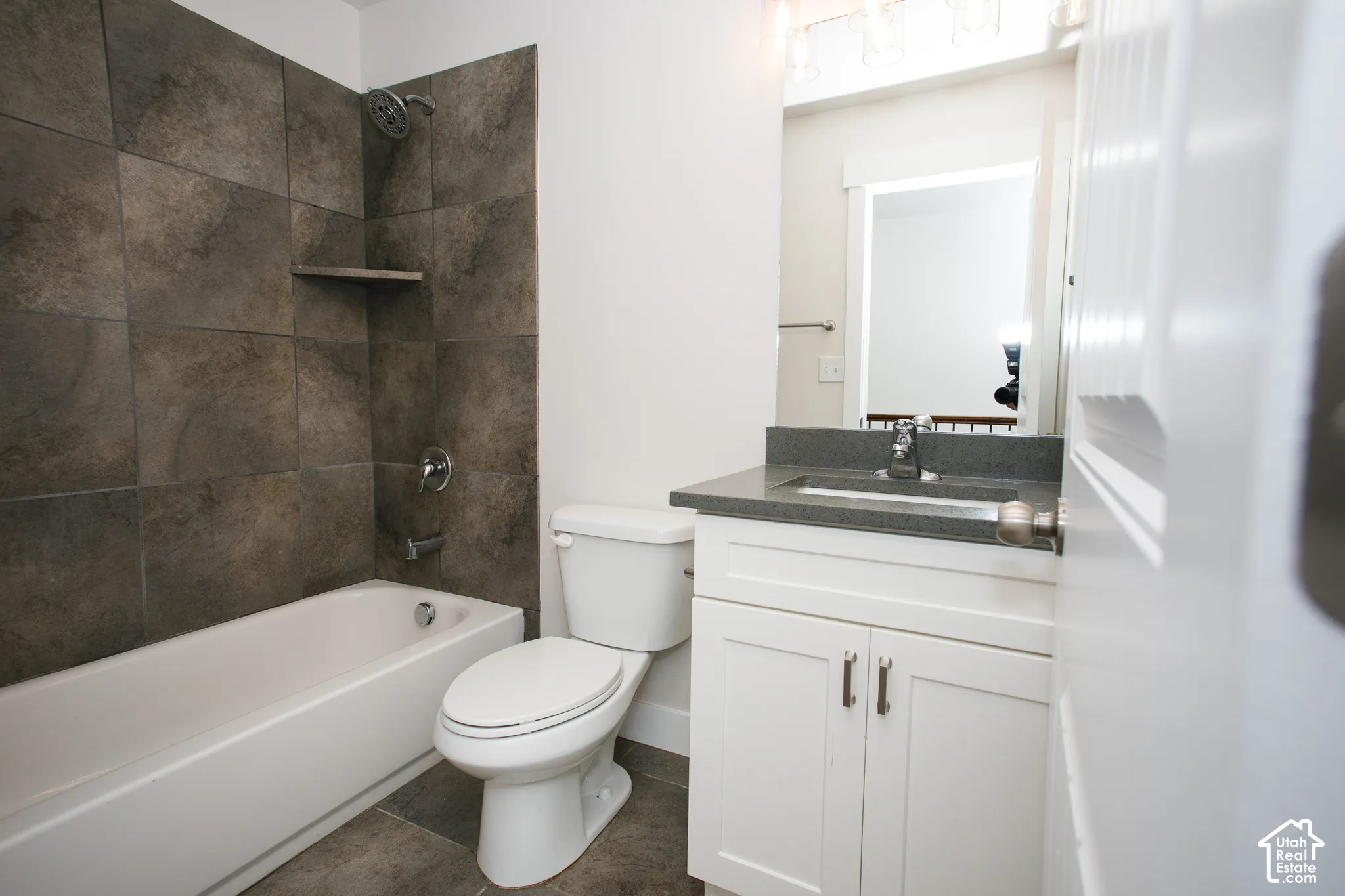 Bathroom featuring a white vanity cabinet with a dark solid-surface countertop, an integrated sink, large-format dark tile tub surround, and a chrome shower fixture