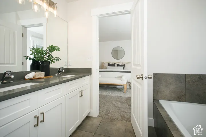 Bathroom featuring a dual-sink vanity with white shaker-style cabinetry, dark stone-finish countertops, polished chrome fixtures, and a soaking tub with dark tile surround
