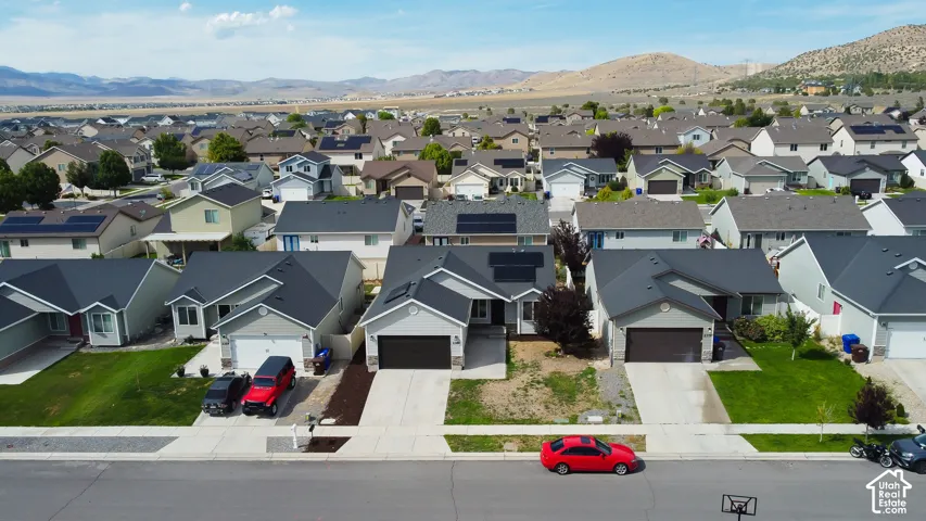 Aerial perspective featuring a residential property with an attached two-car garage, roof-mounted solar panels, concrete driveway, and mountain backdrop