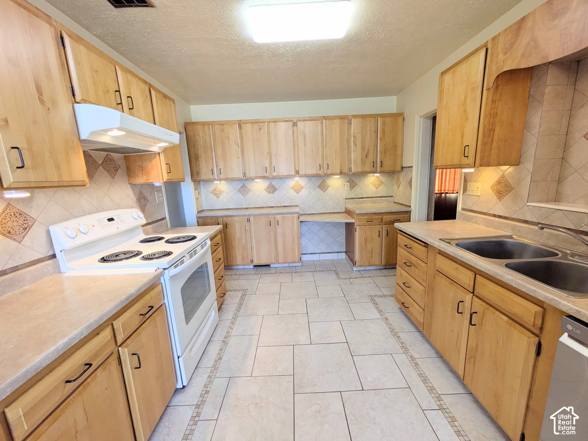 Kitchen featuring light-tone wood cabinetry, tile flooring with mosaic inlay borders, neutral-tone laminate countertops, decorative tile backsplash, and built-in under-cabinet lighting