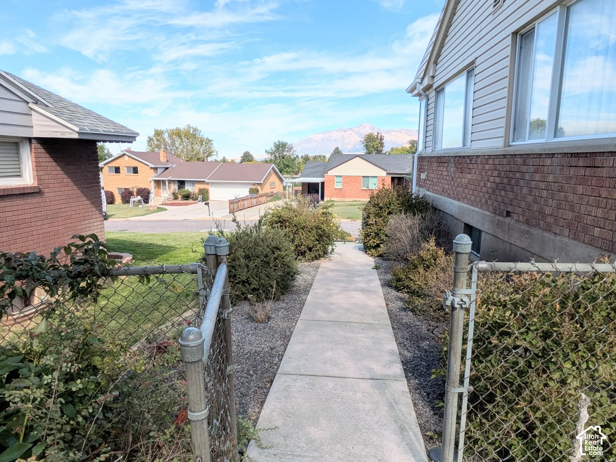 Concrete walkway flanked by chain-link fencing and mature shrubbery, featuring a mountain backdrop