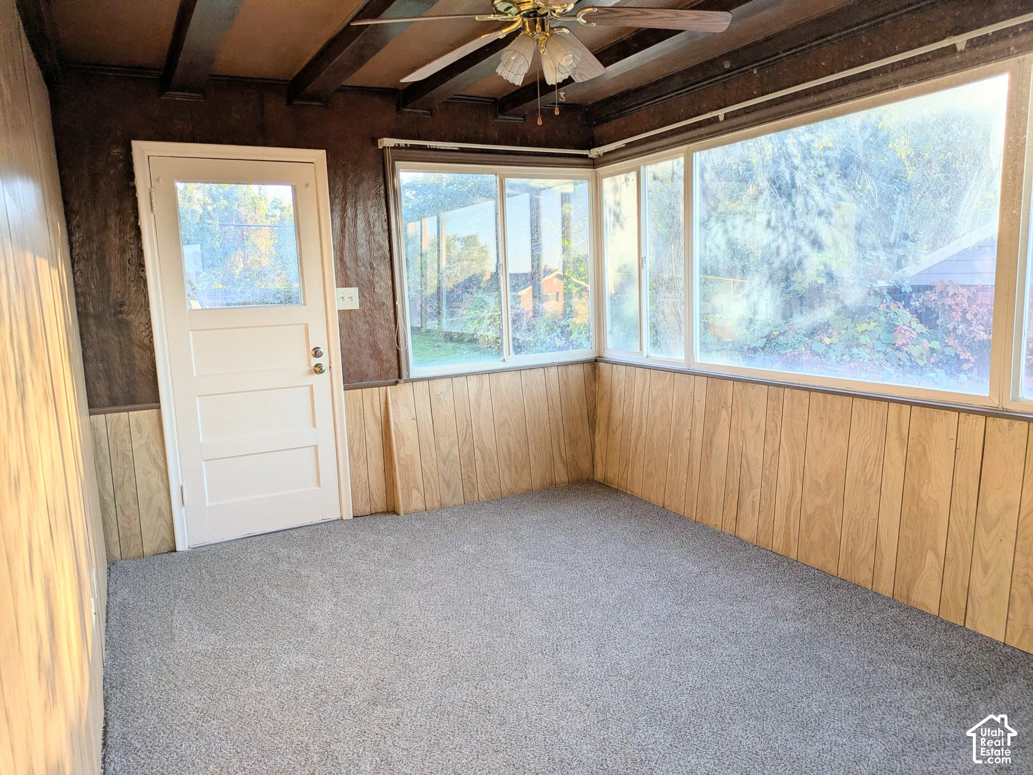 Enclosed space featuring wood paneling, wood-finish ceiling beams, expansive windows, carpet flooring, and an exterior door with a glass inlay