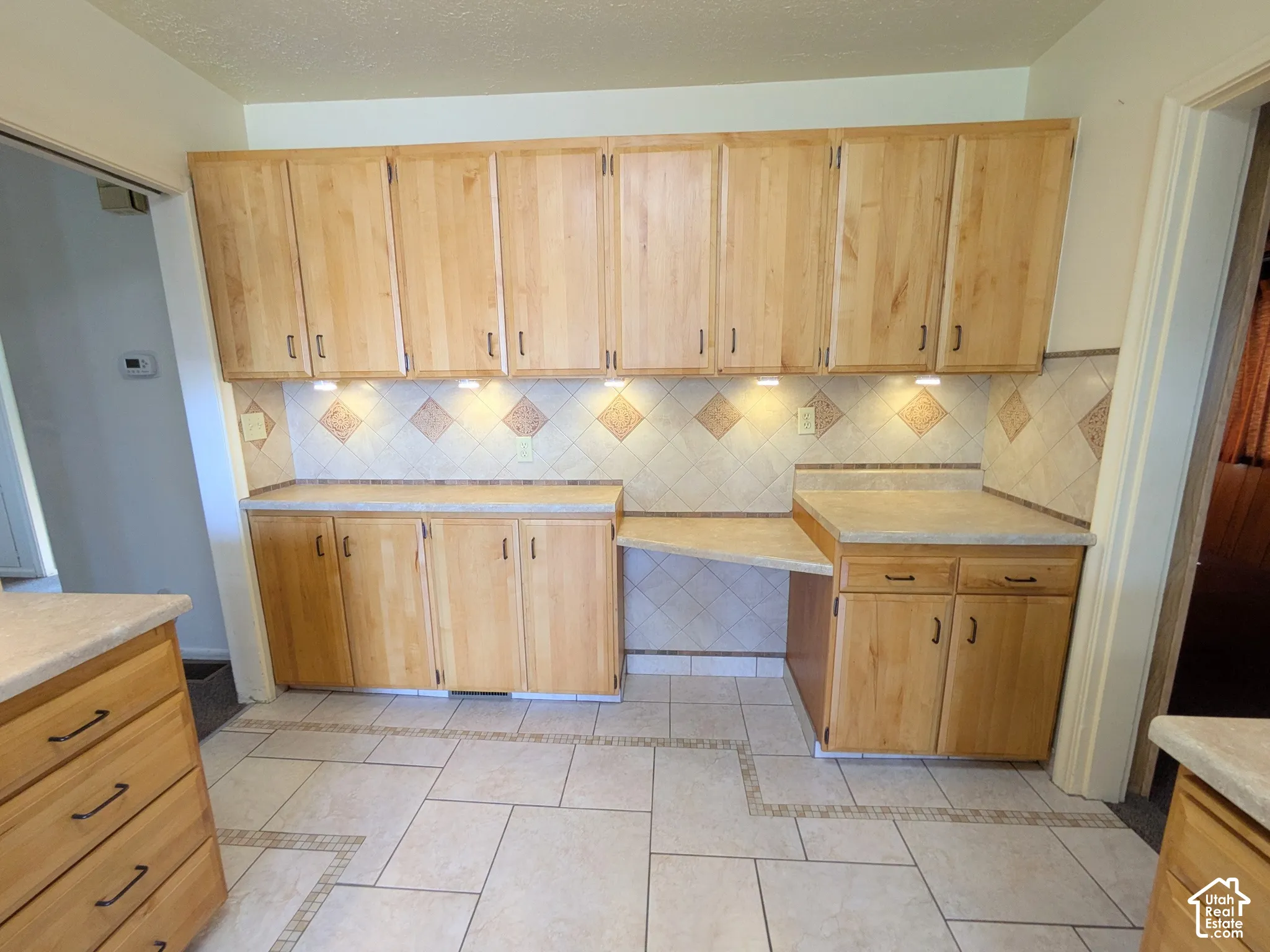 Kitchen cabinetry featuring natural wood-finish doors, light-toned laminate countertops, diamond-pattern tile backsplash with decorative accents, and tiled flooring