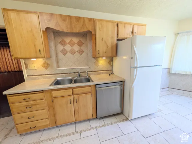 Kitchen featuring natural wood cabinetry, a dual-basin stainless steel sink, tiled backsplash with diamond-pattern detailing, and ceramic tile flooring