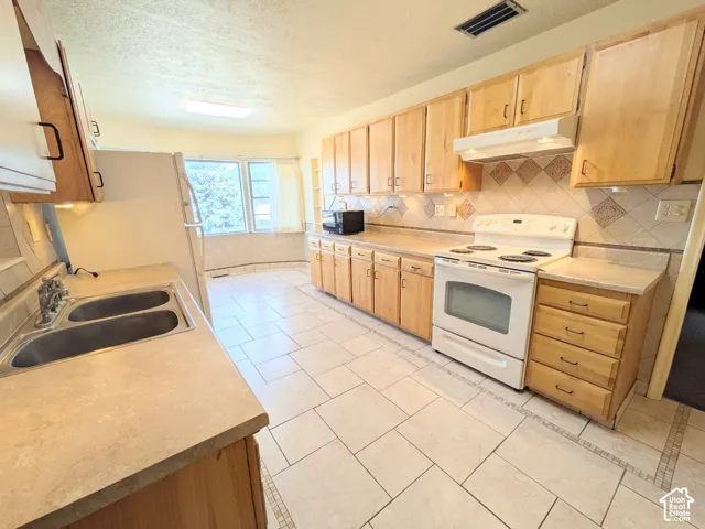 Kitchen featuring light wood cabinetry, white electric range with hood, double-basin stainless steel sink, patterned tile backsplash, and tile flooring