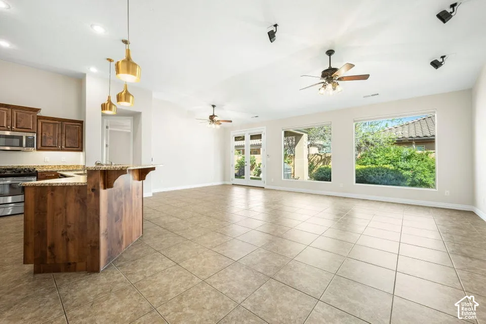 Open-concept interior featuring large-format tile flooring, stained wood cabinetry with a raised breakfast bar, vaulted ceilings, dual ceiling fans, and expansive windows