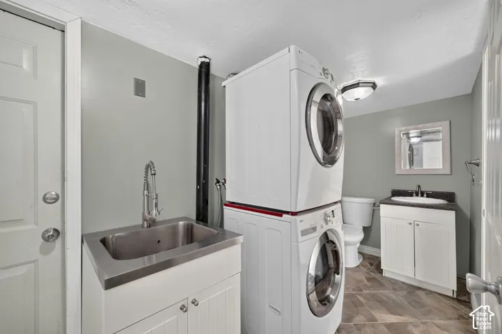 Laundry area featuring a utility sink with a high-arch pull-down faucet, stacked laundry units, tile flooring, and a white vanity with a dark countertop and integrated sink