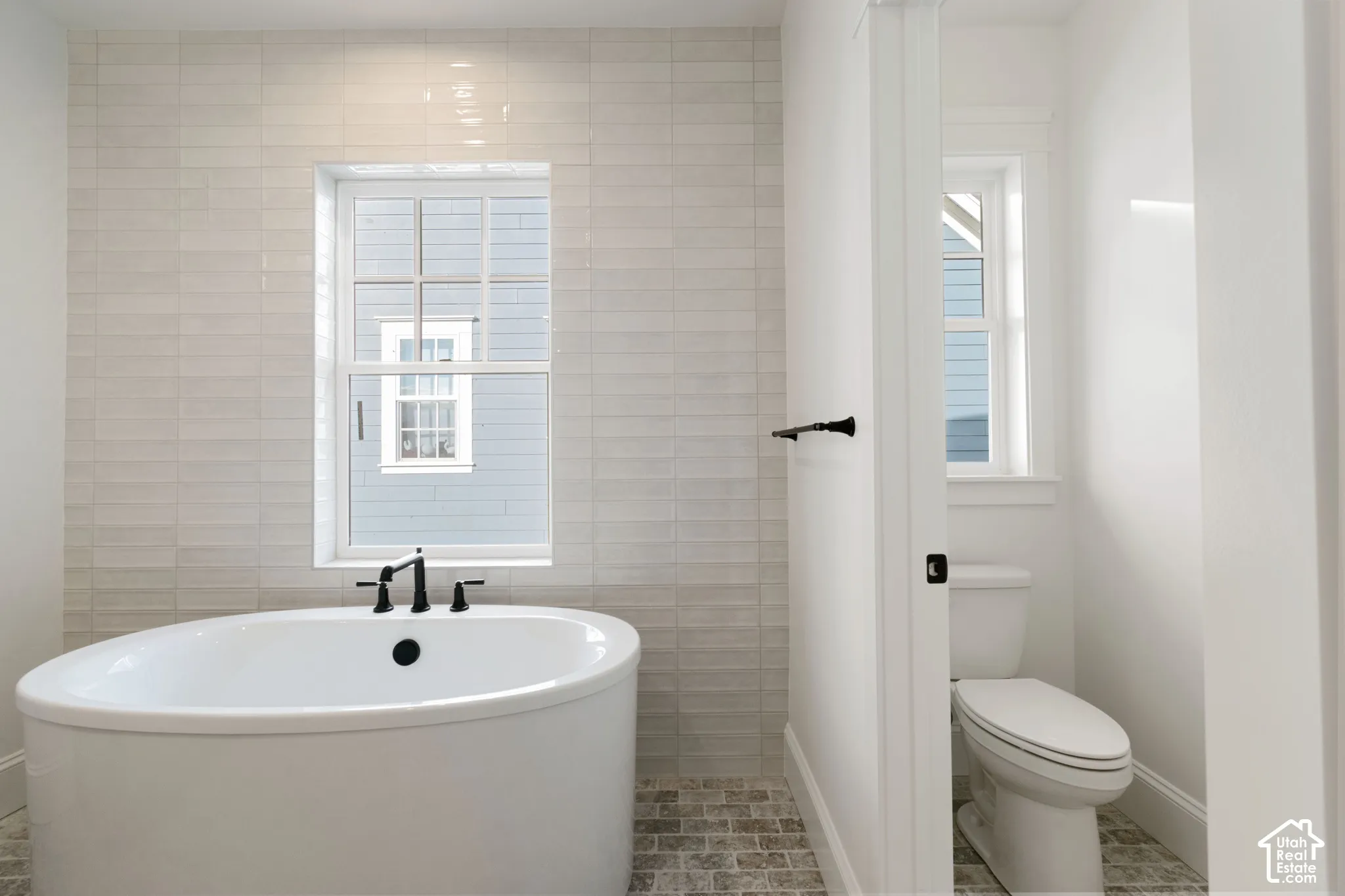 Freestanding soaking tub featuring a matte black deck-mounted faucet, set against a floor-to-ceiling light-toned tile accent wall with a built-in window and stone-finish floor tiling